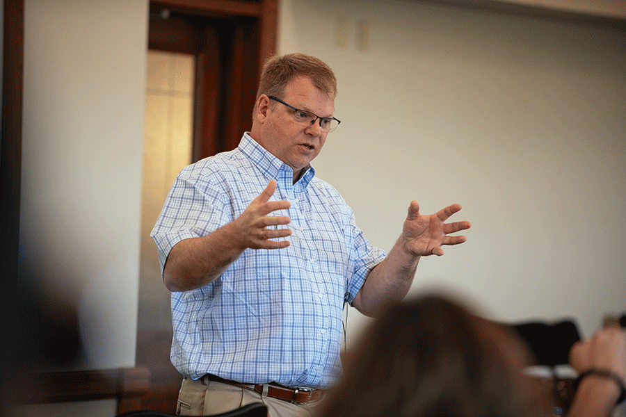 A white middle-aged male with short brown hair stands in a room, talking to a group of individuals blurred in the photo. He wears glasses, a white-and-blue checkered dress shirt, and khaki dress pants. Both of his hands are raised as he talks. 